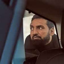 A man with a beard gives a suspicious side-eye look from a car window.